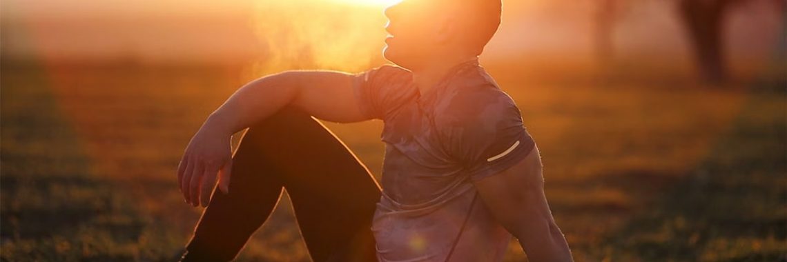 Muscular man sits in rural field at sunset, exhaling visible breath.